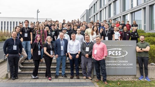 Na zdjęciu grupowym przed nowoczesnym, szarym budynkiem stoi duża grupa ludzi, prawdopodobnie uczestników wydarzenia lub pracowników instytucji. W centrum pierwszego planu znajduje się sześciu mężczyzn i dwie kobiety, którzy wydają się być organizatorami lub przedstawicielami, pozujących przed szarym znakiem z logo PCSS i tekstem "POZNAŃSKIE CENTRUM SUPERKOMPUTEROWO-SIECIOWE". Pozostali uczestnicy stoją na schodach za nimi, uśmiechając się do kamery.