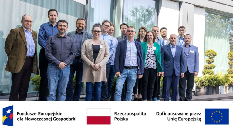 Grupowe zdjęcie przedstawia około 17 osób w różnym wieku, stojących przed nowoczesnym budynkiem. W tle widoczny jest zielony krajobraz. U dołu zdjęcia znajdują się logotypy programów wspierających rozwój gospodarczy: "Fundusze Europejskie dla Nowoczesnej Gospodarki", "Rzeczpospolita Polska" oraz "Dofinansowane przez Unię Europejską".