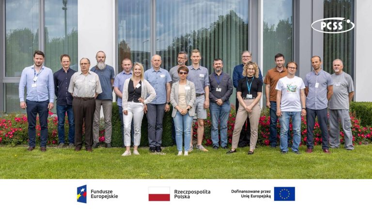 Zdjęcie przedstawia grupę dorosłych osób stojących na trawniku przed nowoczesnym budynkiem z dużymi oknami. Łącznie jest 19 osób, zarówno mężczyzn, jak i kobiet, ubranych w stroje typu biznes casual. W tle widać zielone krzewy i budynek, natomiast na pierwszym planie znajduje się zadbany trawnik i rabata z różowymi kwiatami. Na dole obrazu widoczne są trzy logotypy z tekstem, sugerujące zaangażowanie środków europejskich i polskich.