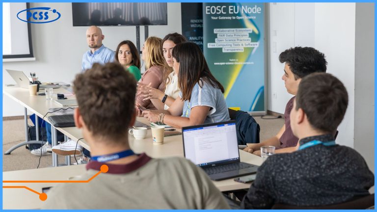 This is a picture taken during the EmpowerHer meeting at PCSS. The image captures a group of people seated around a long table in what appears to be a conference room or classroom setting. In the foreground, the back of a person's head and shoulders are visible, facing away from the viewer. Around the table, several individuals are engaged in discussion or listening intently, with laptops, notebooks, and drinks scattered across the table. A blue and white logo, possibly for "PCSS," is visible in the top left corner of the image.