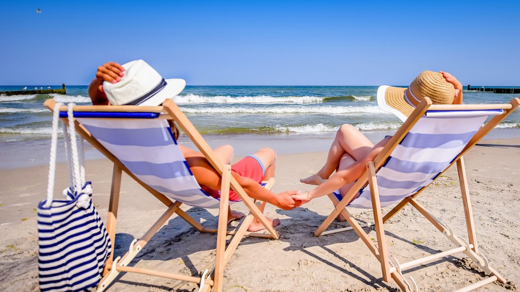 PPAM 2024 Na piaszczystej plaży dwoje ludzi siedzi na leżakach, patrząc na morze. Słońce świeci, a błękitne niebo rozciąga się nad spokojnymi falami. Obok leżaka po lewej stronie wisi torba plażowa w biało-niebieskie paski.