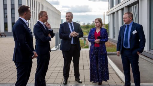 Obraz przedstawia pięć osób w strojach biznesowych, stojących na zewnątrz nowoczesnego budynku i prowadzących rozmowę. Trzech mężczyzn jest ubranych w garnitury, a kobieta ma na sobie elegancki, wzorzysty komplet. W tle widoczne są nowoczesne budynki i zachmurzone niebo.