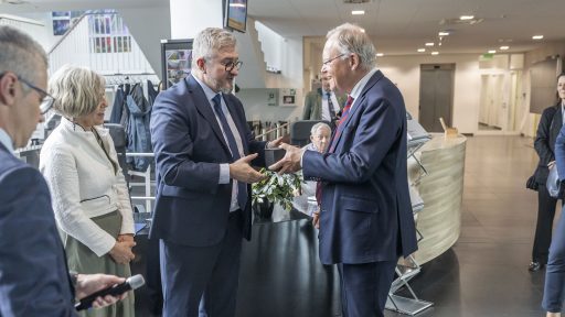 Trzech elegancko ubranych dorosłych uśmiecha się i rozmawia. Kobieta pośrodku trzyma mikrofon i patrzy na mężczyznę po jej lewej stronie. Mężczyzna po prawej stronie również się uśmiecha, patrząc w kierunku kobiety.