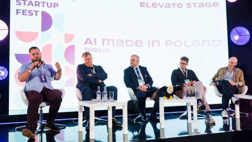 Obraz przedstawia panel dyskusyjny na scenie konferencji, prawdopodobnie Carpathian Startup Fest, sądząc po logo widocznym na ekranie. Na scenie siedzi pięciu mężczyzn, a przed nimi znajdują się słuchacze. Tło sceny jest podświetlone na niebiesko i fioletowo, z różnymi wzorami graficznymi.