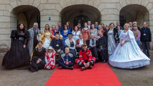 Na zdjęciu widać dużą grupę ludzi, wielu z nich ubranych w historyczne lub eleganckie stroje, stojących i siedzących na czerwonym dywanie przed kamienną fasadą budynku z arkadami. Wśród nich wyróżniają się osoby w długich, balowych sukniach oraz mężczyźni w garniturach i mundurach. Scena wygląda na inscenizację lub wydarzenie tematyczne z udziałem wielu uczestników.