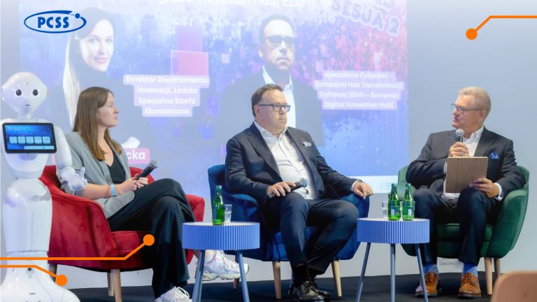 Query successful Trzy osoby dorosłe siedzą na scenie przed dużym ekranem, podczas konferencji. Po lewej stronie, obok kobiety, znajduje się biały robot humanoidalny. Na stolikach przed panelistami stoją butelki z wodą i szklanki.