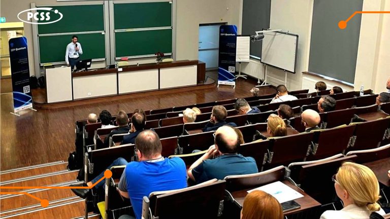 Query successful Prelegent przemawia do publiczności w auli. Mężczyzna w jasnoniebieskiej koszuli stoi za długim stołem na przodzie sali, zwrócony w stronę rzędów siedzących słuchaczy. Za prelegentem widoczne są dwie duże zielone tablice, a w lewym górnym rogu obrazu znajduje się logo "PCSS".