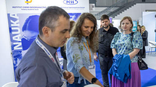 Na zdjęciu widać grupę osób stojących na targach lub konferencji naukowej przy stoisku Instytutu Chemii Bioorganicznej Polskiej Akademii Nauk. W tle znajduje się niebiesko-białe stoisko z dużym napisem „NAUKA” oraz grafiką przedstawiającą osobę w laboratorium. Uczestnicy rozmawiają ze sobą, a jedna z kobiet trzyma w ręku niebieską kurtkę.