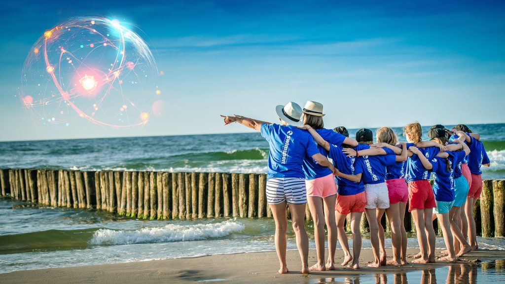 Zdjęcie przedstawia grupę dzieci i dorosłych, prawdopodobnie opiekunów, stojących tyłem do obiektywu na piaszczystej plaży. Patrzą oni na morze, a na horyzoncie, po lewej stronie, unosi się świetlista kula, przypominająca schemat atomu lub sieci połączeń. Kompozycja zdjęcia sugeruje beztroskie wakacje połączone z futurystycznym lub naukowym motywem.