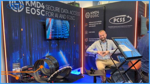 Zdjęcie przedstawia stoisko konferencyjne, prawdopodobnie na konferencji ISC25 w Hamburgu. W centrum uwagi znajduje się mężczyzna z brodą, ubrany w jasną koszulę i beżowe spodnie, siedzący na krześle i patrzący wprost w obiektyw. W tle widać banery z napisami "KMD4 SECURE DATA ACCESS EOSC FOR AI AND EOSC" oraz "PCSS Poznańskie Centrum Superkomputerowo-Sieciowe”, co sugeruje, że stoisko dotyczy dostępu do danych w kontekście sztucznej inteligencji i EOSC. Na stole przed mężczyzną stoi mały tor wyścigowy z samochodzikami, a na monitorach wyświetlają się grafiki związane z danymi i superkomputerami.