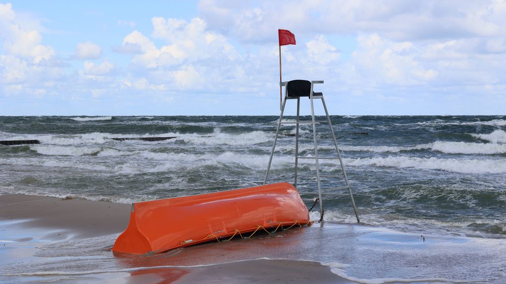 Flaga ratunkowa wskazuje na wzburzone morze. Na plaży stoi puste krzesło ratownika, a obok niego leży odwrócona, pomarańczowa łódź. To zdjęcie oddaje nastrój ponurego, chłodnego i nieprzyjaznego dnia na brzegu morza.