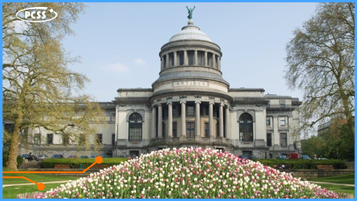 Duży, neoklasycystyczny budynek z kopułą, frontowymi kolumnami i napisem „ARTIBVS” widnieje w centrum kadru, a na jego szczycie umieszczono rzeźbę. Na pierwszym planie znajduje się duży, usypany kopiec pokryty kwitnącymi, biało-różowymi tulipanami. Po obu stronach budynku rosną drzewa z jasnymi liśćmi, a pod niebem widać logo PCSS.