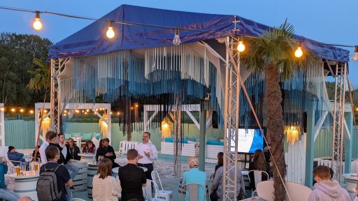 Mężczyzna w białej koszuli i czarnych spodniach stoi pod zadaszeniem, prowadząc prezentację dla grupy ludzi, którzy siedzą na krzesłach i drewnianych skrzyniach. W tle znajduje się piaszczysta plaża, na której rozstawiono błękitne sofy i poduszki, a oświetlenie tworzą żarówki podwieszone pod zadaszeniem. Całość odbywa się wieczorem w otoczeniu palm i drewnianej zabudowy.