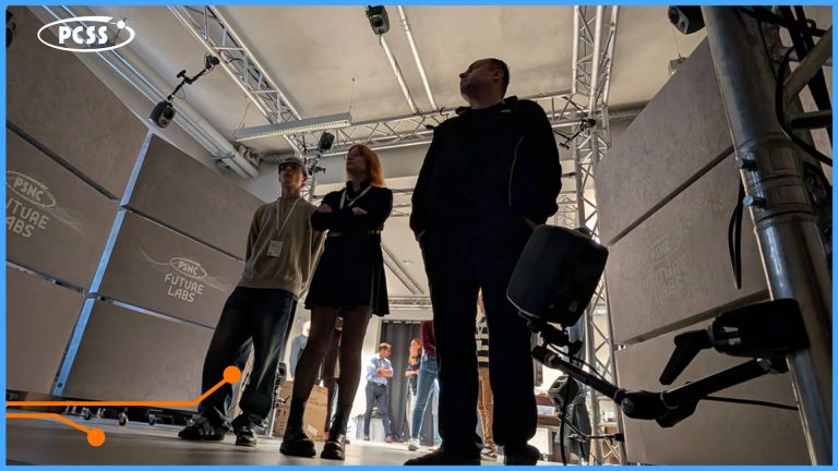 Trzy osoby stoją pod dużą sceną lub instalacją, której częścią są duże, szare panele z napisami PSNC FUTURE LABS. Dwóch mężczyzn i kobieta patrzą w górę i w prawo, a ich sylwetki są przedstawione z perspektywy z dołu. Scena i widownia, w tym więcej osób stojących dalej, są oświetlone, a u góry widoczne jest logo PCSS.