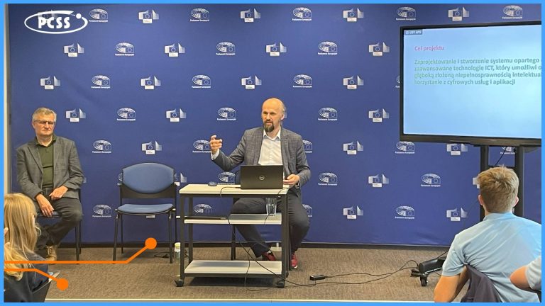 Konferencja lub prezentacja odbywa się w sali, gdzie na ścianie wiszą powtarzające się logotypy, w tym logo PCSS oraz unijne gwiazdy. Na pierwszym planie, za stołem z laptopem, stoi mężczyzna w garniturze i z brodą, który gestykuluje podczas przemawiania do audytorium. Obok niego po lewej siedzi drugi mężczyzna w okularach i marynarce, a po prawej stronie kadru widoczna jest część ekranu z tekstem w języku polskim oraz fragmenty słuchaczy.