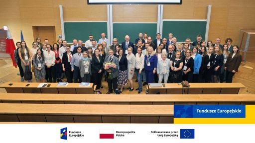 Ta szeroka fotografia w auli przedstawia dużą grupę osób — prawdopodobnie uczestników konferencji lub szkolenia — stojących w rzędach. Uczestnicy, głównie dorośli w strojach biznesowych lub półformalnych, uśmiechają się i patrzą w kierunku aparatu, a w centralnej części jedna osoba trzyma duży bukiet kwiatów. Na dole zdjęcia umieszczono paski z logotypami i flagami: Fundusze Europejskie, flaga Rzeczypospolitej Polskiej oraz informacja "Dofinansowane przez Unię Europejską".