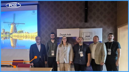 Zdjęcie przedstawia sześcioosobową grupę dorosłych osób pozujących do zdjęcia w sali konferencyjnej. Grupa stoi przed bannerem z logotypami DARIAH-HUB oraz PAN (Polskiej Akademii Nauk), na tle którego widać również logo PCSS na ekranie projektora. Ekran za nimi wyświetla relaksujące zdjęcie holenderskich wiatraków nad wodą, co sugeruje przerwę lub zakończenie spotkania.