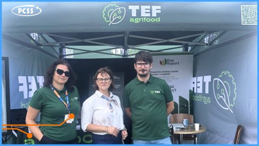 Zdjęcie przedstawia trzy osoby – dwie kobiety i mężczyznę – stojące pod namiotem promocyjnym z logotypem PCSS i TEF agrifood na targach lub wydarzeniu plenerowym. Osoby te, ubrane w koszulki polo z logo TEF lub odzież biznesową, są prawdopodobnie pracownikami lub reprezentantami projektu. Za nimi widoczne są materiały informacyjne, w tym baner z hasłem "Risk Raport" oraz tekst o eksperckiej analizie ryzyka maszyn i urządzeń.