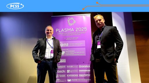 Zdjęcie przedstawia dwóch mężczyzn w ciemnych garniturach stojących obok siebie na tle dużej, fioletowej roll-upy z informacjami o konferencji. Roll-up dotyczy "PLASMA 2025", czyli "International Conference on Research & Application of Plasmas", która ma odbyć się w Warszawie. Mężczyźni, z identyfikatorami na szyjach, uśmiechają się do obiektywu, a scena sugeruje, że są prelegentami, uczestnikami lub organizatorami tego naukowego wydarzenia.