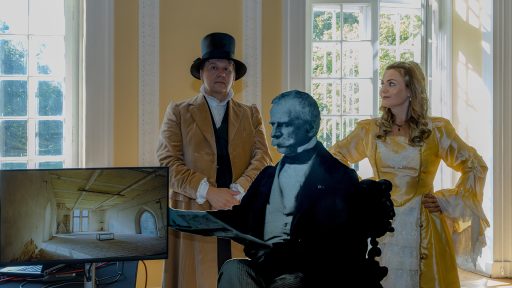 Zdjęcie przedstawia dwie osoby w strojach z epoki, stojące w jasnym pokoju z wysokimi oknami. Osoba po lewej ma na sobie brązowy kostium i cylinder, a po prawej jasną, żółtą suknię. Pomiędzy nimi znajduje się naturalnej wielkości wycięta postać mężczyzny ze zdjęcia czarno-białego, a po lewej stronie monitor wyświetla wnętrze historycznego pomieszczenia.