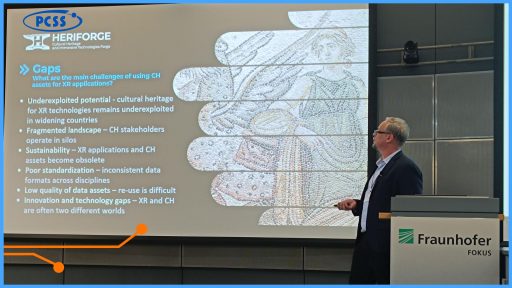 Zdjęcie przedstawia prelegenta w garniturze stojącego po prawej stronie dużego ekranu projekcyjnego w sali konferencyjnej. Na ekranie widać tytuł "Gaps" i listę wyzwań związanych z wykorzystaniem dóbr dziedzictwa kulturowego (CH) w aplikacjach XR, obok której wyświetlany jest duży, ozdobny motyw mozaiki. W dolnym prawym rogu ekranu i sali widoczne jest logo Fraunhofer FOKUS.