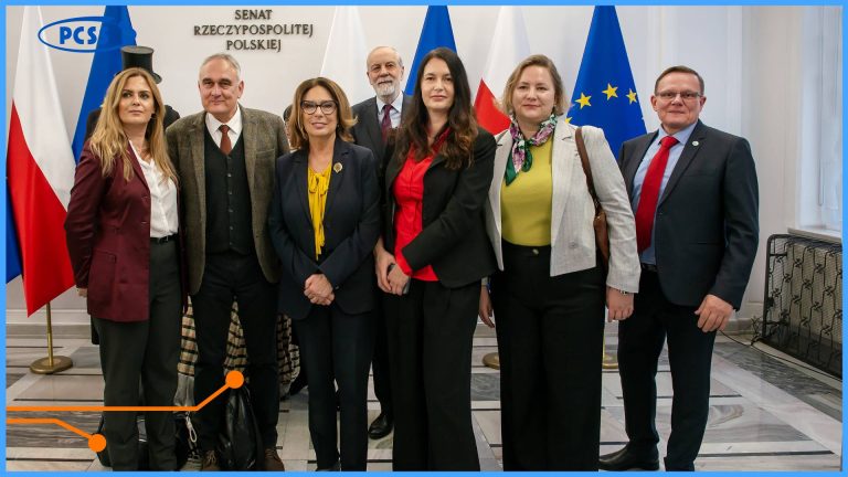 Grupowe zdjęcie siedmiu osób, w tym trzech mężczyzn i czterech kobiet, wykonane w holu Senatu Rzeczypospolitej Polskiej, na co wskazuje napis na ścianie oraz flagi polskie i unijne. W centrum widoczna jest Marszałek Senatu, Małgorzata Kidawa-Błońska, w żółtej bluzce i ciemnym garniturze, otoczona przez przedstawicieli Instytutu Chemii Bioorganicznej PAN oraz PCSS. Osoby stoją w rzędzie, uśmiechnięte, ubrane w stroje biznesowe i półformalne, co sugeruje oficjalne spotkanie w ramach otwarcia wystawy o pracy organicznej.