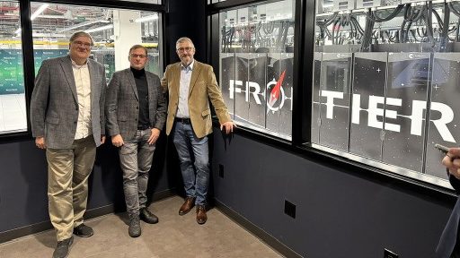 Na zdjęciu widać trzech mężczyzn w eleganckich strojach biznesowych, stojących przed dużą przeszkloną ścianą. Za szybą znajduje się serwerownia, w której widoczne są szafy z dużym logo "FRONTIER". Mężczyźni patrzą w kierunku aparatu, a przestrzeń w pomieszczeniu jest wykończona w ciemnych kolorach, co kontrastuje z jasnym oświetleniem serwerowni.