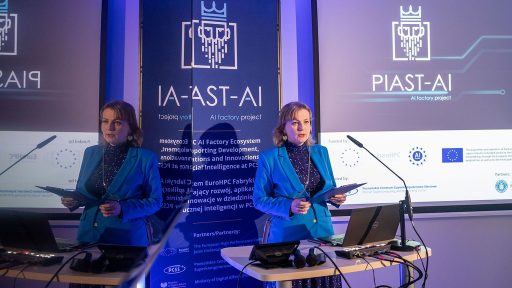Zdjęcie przedstawia prelegentkę stojącą przy pulpicie w sali konferencyjnej, podczas prezentacji związanej z projektem technologicznym. Kobieta ubrana jest w niebieską marynarkę i trzyma kartki w dłoniach, a obok niej znajdują się mikrofon, laptop i monitor odsłuchowy. W tle widoczne są duże grafiki projektowe z logotypem "PIAST-AI" oraz informacje o finansowaniu przez EuroHPC i Partnerstwach, co wskazuje na prezentację dotyczącą rozwoju sztucznej inteligencji.