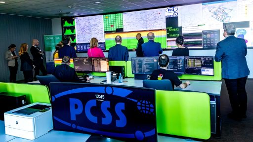 Zdjęcie przedstawia wnętrze nowoczesnego centrum monitorowania, należącego do PCSS. W centralnej części pomieszczenia duży ekran za stołem z monitorami wyświetla schematy sieci i dane w czasie rzeczywistym, a przed nim, w głębi, stoi grupa formalnie ubranych osób (prawdopodobnie delegacja). Na pierwszym planie widoczny jest monitor z dużym logo PCSS, a pracownicy siedzą przy biurkach z jaskrawozielonymi przegrodami, monitorując systemy.