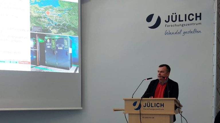 Mężczyzna przemawia zza mównicy z logo Forschungszentrum Jülich podczas oficjalnej prezentacji. W tle widoczny jest ekran wyświetlający mapę sieci połączeń oraz zdjęcia nowoczesnych szaf serwerowych z oznaczeniami projektów kwantowych. Na ścianie obok prelegenta znajduje się duże logo ośrodka badawczego z hasłem „Wandel gestalten”.