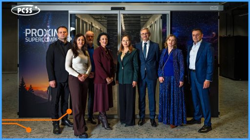 Zdjęcie przedstawia grupę ośmiu dorosłych osób – czterech kobiet i czterech mężczyzn – pozujących na tle dużej przeszklonej szafy serwerowej z napisem PCSS PROXIMA SUPERCOMPUTING. Wszyscy są ubrani w stroje formalne lub półformalne, z dwoma mężczyznami po bokach w niebieskich garniturach i dwiema kobietami w środku, z których jedna ma ciemnozieloną garsonkę, a druga długą, błyszczącą niebieską suknię. Kompozycja zdjęcia jest formalna i prawdopodobnie dokumentuje wizytę ważnych osobistości w centrum superkomputerowym.