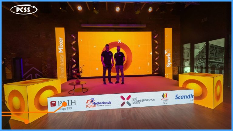 Two PCSS representatives in dark shirts stand smiling on a vibrant event stage at Spark Demo Day in Łódź, with large yellow cubic displays, orange Spark and Mixer branding on screens and backdrops, and sponsor logos including PFR, PARP, and Scandio.