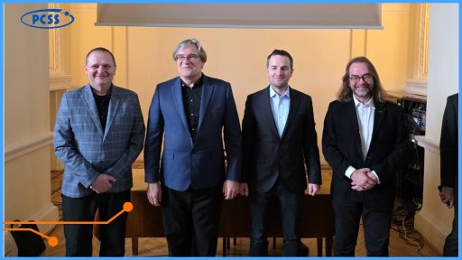 Czterech mężczyzn w garniturach i marynarkach stoi obok siebie, uśmiechając się do zdjęcia w jasnym pomieszczeniu. W lewym górnym rogu widoczne jest logo PCSS, a w tle znajduje się ekran projekcyjny oraz sprzęt elektroniczny. Postacie ustawione są w rzędzie na tle jasnej ściany i drewnianego stołu.