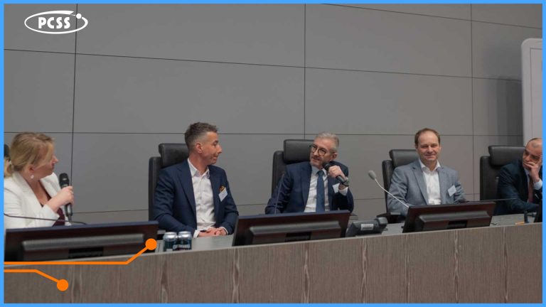 Zdjęcie przedstawia panel dyskusyjny z udziałem pięciu osób siedzących przy długim stole w nowoczesnej sali konferencyjnej. Pośrodku widoczny jest Robert Pękał z PCSS, który wypowiada się do mikrofonu, podczas gdy pozostali uczestnicy słuchają lub prowadzą rozmowę. W lewym górnym rogu grafiki umieszczono logotyp PCSS, a całość oprawiona jest w niebieską ramkę z pomarańczowym elementem graficznym na dole.