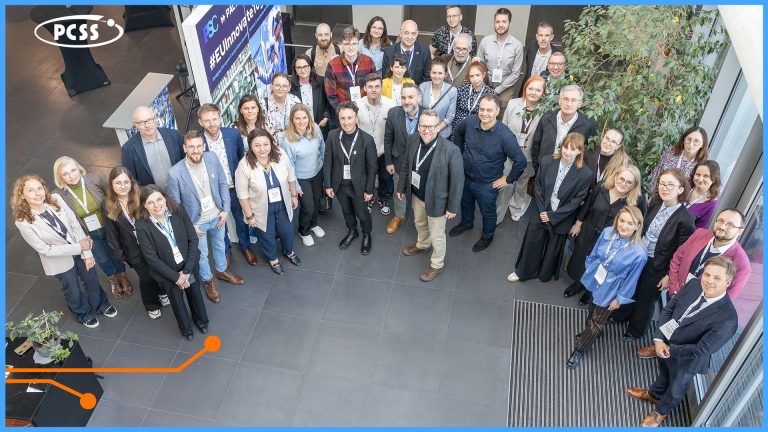 Query successful Zdjęcie grupowe wykonane z góry przedstawia kilkudziesięciu uczestników wydarzenia w nowoczesnym, przestronnym wnętrzu z logo PCSS w lewym górnym rogu. Osoby o różnych profesjach są uśmiechnięte, ubrane w stroje biznesowe i stoją na tle przeszklonej ściany oraz zielonych roślin doniczkowych. Kompozycja zdjęcia podkreśla wspólnotowy i profesjonalny charakter spotkania.