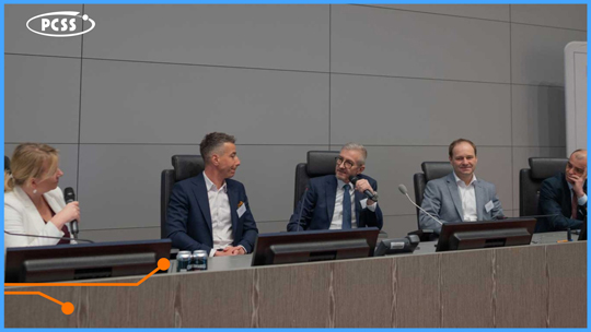 Zdjęcie przedstawia panel dyskusyjny z udziałem pięciu osób siedzących przy długim stole w nowoczesnej sali konferencyjnej. Pośrodku widoczny jest Robert Pękał z PCSS, który wypowiada się do mikrofonu, podczas gdy pozostali uczestnicy słuchają lub prowadzą rozmowę. W lewym górnym rogu grafiki umieszczono logotyp PCSS, a całość oprawiona jest w niebieską ramkę z pomarańczowym elementem graficznym na dole.