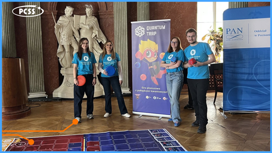 Czworo uśmiechniętych animatorów w niebieskich koszulkach z logo PCSS pozuje w zabytkowym wnętrzu obok wielkoformatowej gry planszowej rozłożonej na podłodze. W tle widoczny jest pionowy baner promujący grę „Quantum Trap” oraz klasyczne rzeźby i niebieska ścianka z logo Polskiej Akademii Nauk. Całość przedstawia stoisko edukacyjne przygotowane na Poznański Festiwal Nauki i Sztuki.