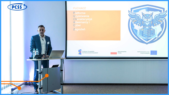 Mężczyzna w garniturze stoi przy mównicy z laptopem, prowadząc prezentację w jasnej sali konferencyjnej. Na dużym ekranie za nim widoczne jest logo projektu "PUCHACZ" (Platforma Uzyskiwania Charakterystyk Adwersarzy i Cyber Zagrożeń) w formie tarczy z sową oraz logotypy Funduszy Europejskich, Rzeczypospolitej Polskiej i Unii Europejskiej. W lewym górnym rogu zdjęcia umieszczono logo PCSS.