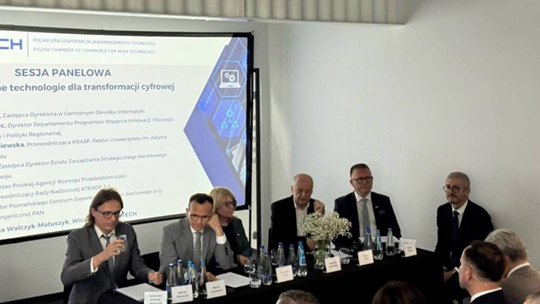 Fotografia przedstawia sześcioosobowy panel dyskusyjny siedzący przy długim, ciemnym stole podczas konferencji zatytułowanej „SESJA PANELOWA”. Po lewej stronie widoczny jest duży ekran projekcyjny z polskim tekstem dotyczącym technologii dla transformacji cyfrowej oraz logo PCSS w prawym górnym rogu kadru. Uczestnicy w eleganckich strojach biznesowych siedzą przed tabliczkami z nazwiskami, butelkami wody oraz centralnie ustawionym bukietem białych kwiatów.