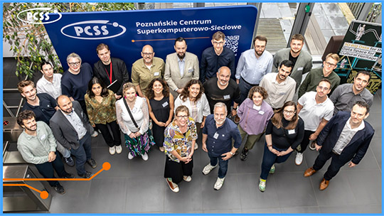 Zdjęcie grupowe uczestników forum technologicznego w holu budynku PCSS. Ponad dwadzieścia osób stoi przed dużą, niebieską ścianką konferencyjną z logo i pełną nazwą „Poznańskie Centrum Superkomputerowo-Sieciowe”. Wszyscy patrzą w stronę obiektywu, który uwiecznił ich z lekkiego podwyższenia.