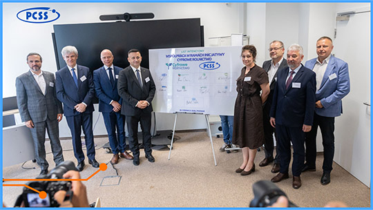 Fotografia przedstawia grupę ośmiu osób w strojach biznesowych, stojących w biurze PCSS po obu stronach dużej, białej tablicy z podpisanym listem intencyjnym dotyczącym współpracy w ramach inicjatywy „Cyfrowe Rolnictwo”. Na tablicy widoczne są logotypy partnerów oraz odręczne podpisy, a w lewym górnym rogu kadru umieszczono logo PCSS. Zdjęcie zostało wykonane z perspektywy fotografa, którego aparat jest częściowo widoczny na pierwszym planie.