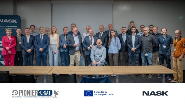 Grupa osób pozuje do zdjęcia grupowego w sali konferencyjnej przed pustym ekranem projekcyjnym. W dolnej części grafiki widoczne są logotypy PIONIER-Q-SAT, Unii Europejskiej oraz NASK. Na pierwszym planie stoi długi, drewniany stół konferencyjny.