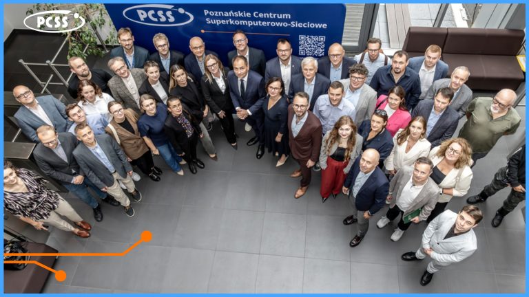 Zdjęcie grupowe wykonane z góry przedstawia kilkadziesiąt osób w strojach biznesowych, stojących w nowoczesnym holu przed niebieską ścianką PCSS. Uczestnicy są ustawieni w półokręgu, patrząc w kierunku obiektywu. Tło stanowi baner z logiem PCSS i pełną nazwą: „Poznańskie Centrum Superkomputerowo-Sieciowe”.