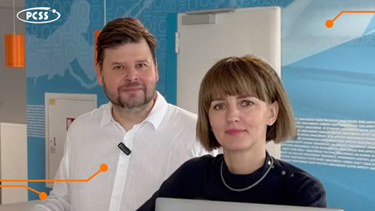 Kobieta i mężczyzna pozują razem w nowoczesnym biurze PCSS. Mężczyzna po lewej ma ciemny zarost i białą koszulę z przypiętym mikrofonem krawatowym, a stojąca obok kobieta ma krótkie, ciemne włosy z grzywką i ciemną bluzkę. W tle widoczne są niebieskie ściany z grafikami oraz logo PCSS w lewym górnym rogu.